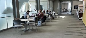 A long view of the common area for the Purdue OWL. Bistro tables where sessions happen, a glass walled conference room beyond, and a meeting space at the end of the all.